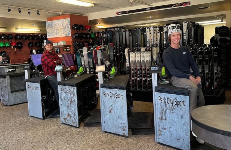 Interior of Park City Sport Ski and Snowboard Rental Shop with two smiling employees sitting on the set up benches with skis in the background