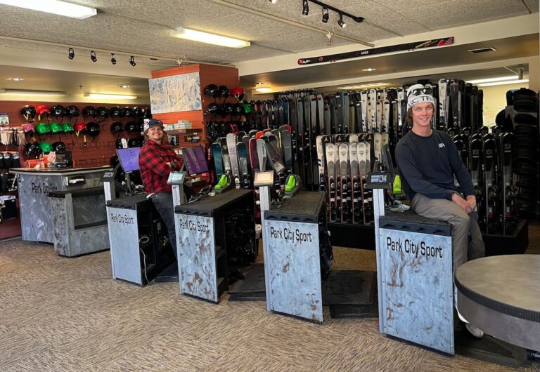 Interior of Park City Sport Ski and Snowboard Rental Shop with two smiling employees sitting on the set up benches with skis in the background